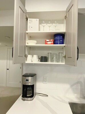 A kitchen with a coffee maker on the counter, a cabinet with various items including a box, cups, and a container.