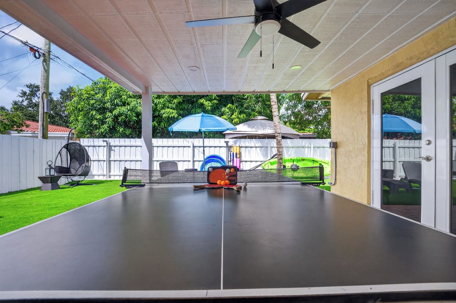 A covered outdoor area with a ping pong table, a ceiling fan, and a white fence in the background.