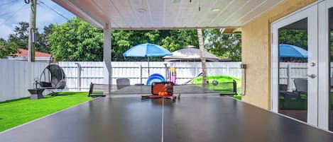 A covered outdoor area with a ping pong table, a ceiling fan, and a white fence in the background.