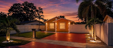 A residential house with a red driveway, a white fence, and a palm tree.