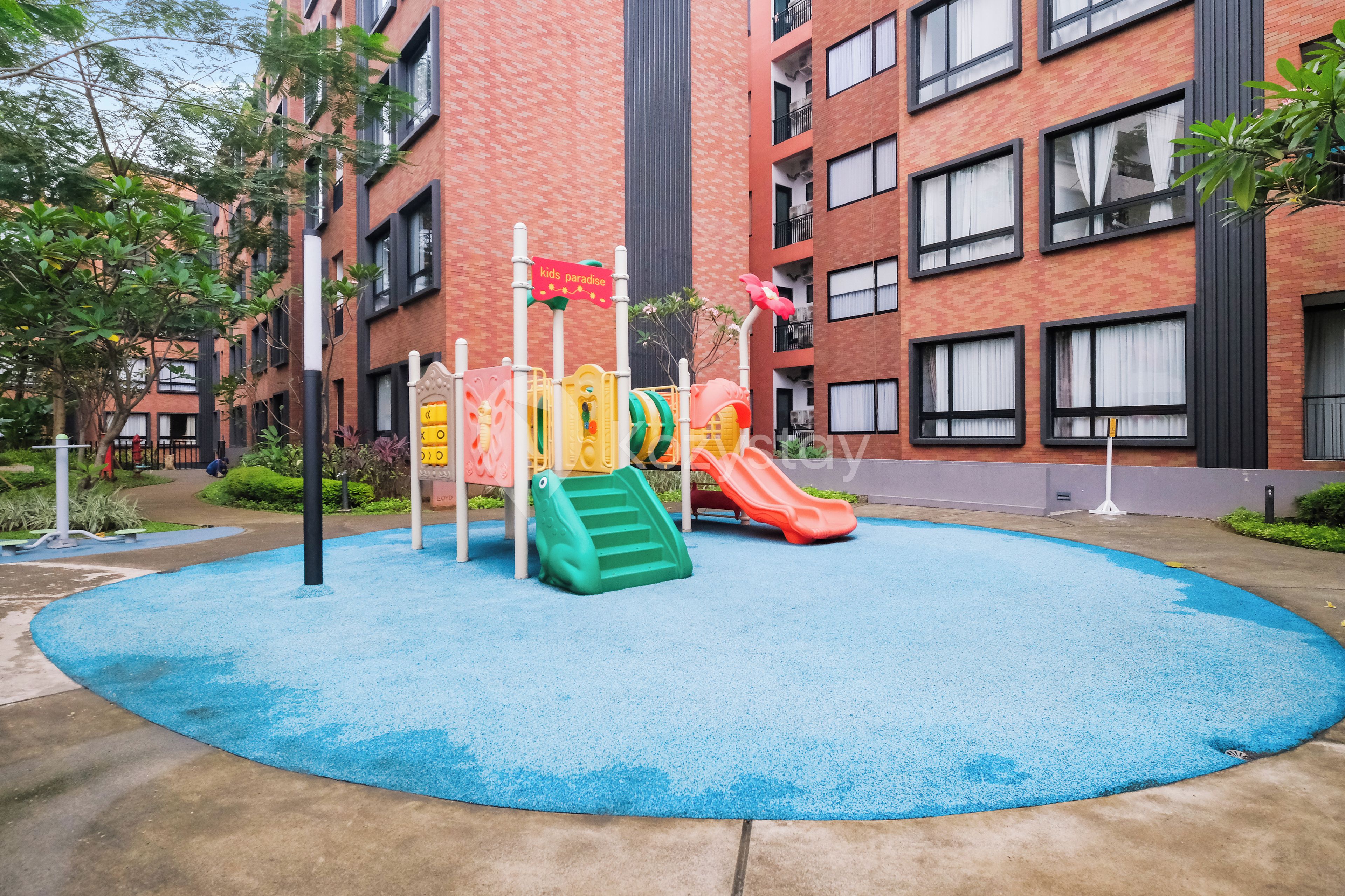 A playground with slides and climbing structures surrounded by a blue play area, set against a brick building with multiple windows.
