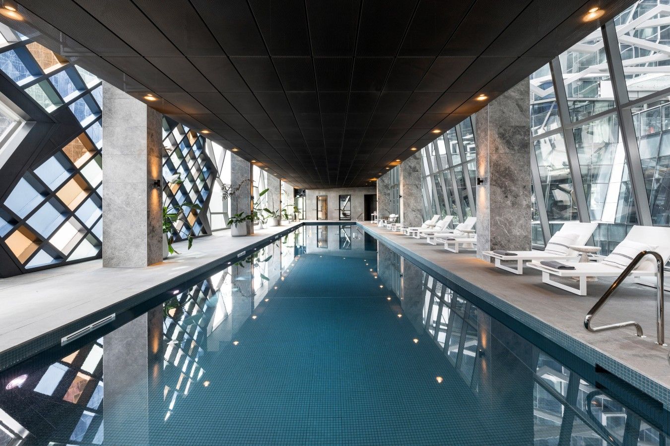 An indoor swimming pool with clear glass walls, lounge chairs, and a modern architectural design.