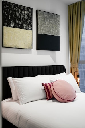 Un lit avec des oreillers blancs et roses, une tête de lit noire et deux tableaux abstraits accrochés au mur.