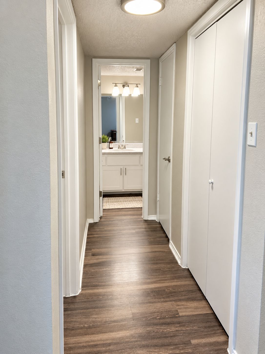 A hallway leading to a bathroom with a vanity, mirror, and lighting fixtures.