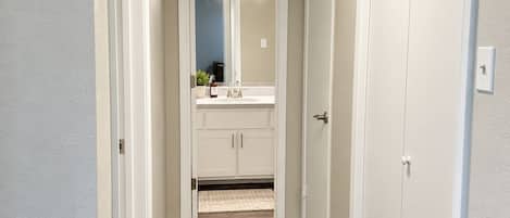 A hallway leading to a bathroom with a vanity, mirror, and lighting fixtures.