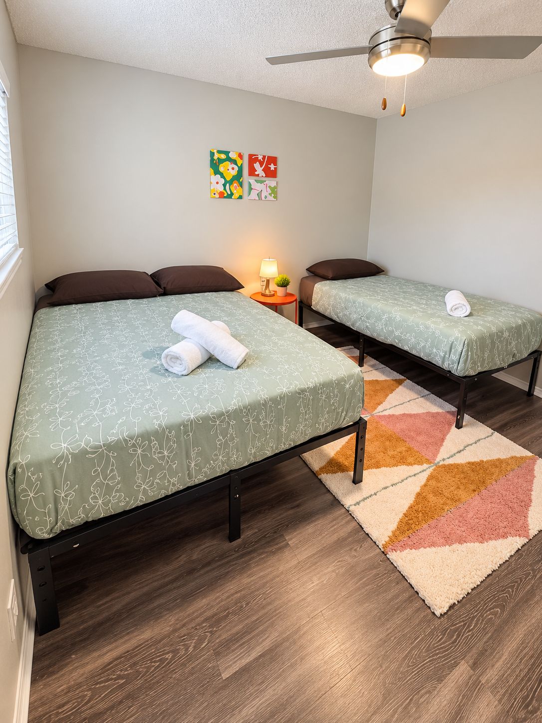 Habitación de hotel con dos camas, un ventilador de techo, una mesita con lámpara y una alfombra colorida.