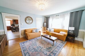 Ein Wohnzimmer mit Sofa, Couchtisch und Teppich. Im Hintergrund ist ein Essbereich zu sehen.