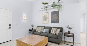A modern living room with a grey sofa, patterned cushions, a woven coffee table, and wall-mounted shelves with plants. - Pinetree Palace - 4BR Cozy Home (Panama City Beach)