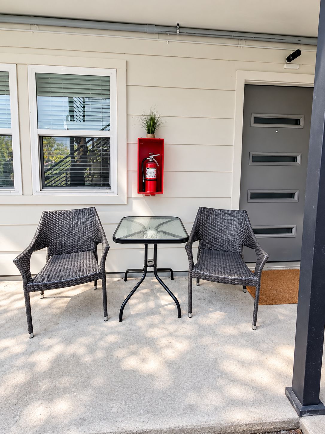 A patio area with two wicker chairs and a glass table, a red fire extinguisher mounted on the wall, and a gray door with a window above it.