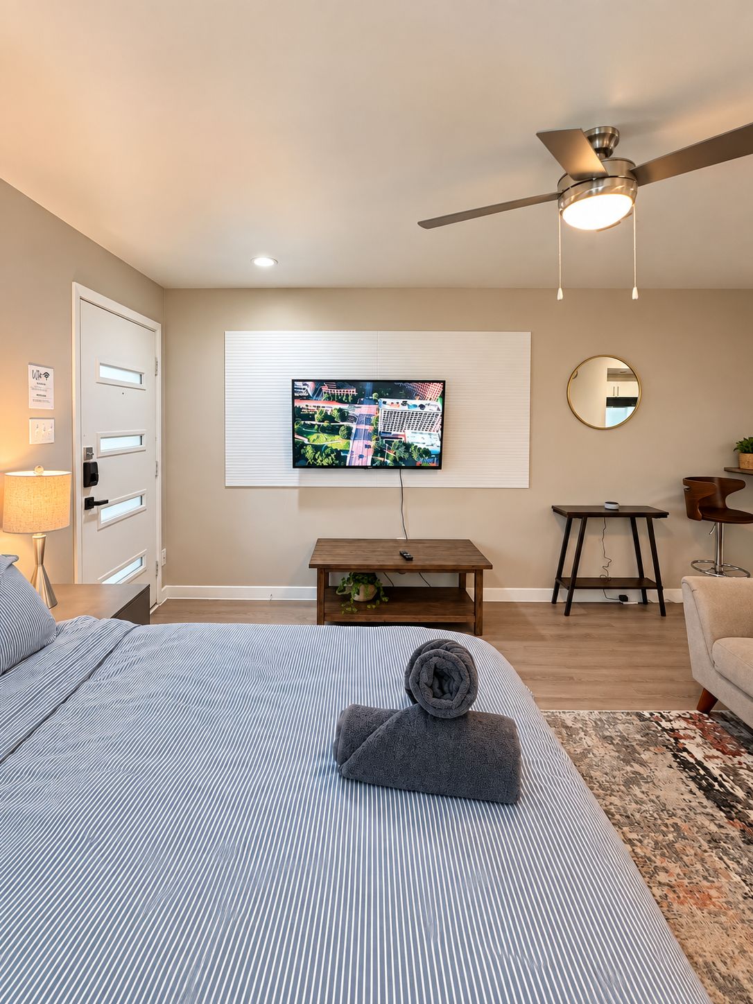 A hotel room with a bed, a television mounted on the wall, a ceiling fan, a small table with a plant, and a round mirror.