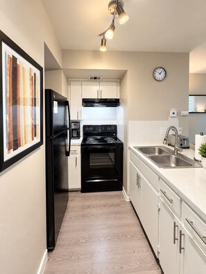 Una cocina moderna con un refrigerador negro, un lavamanos de acero inoxidable y una estufa. Hay un reloj en la pared y un cuadro enmarcado.