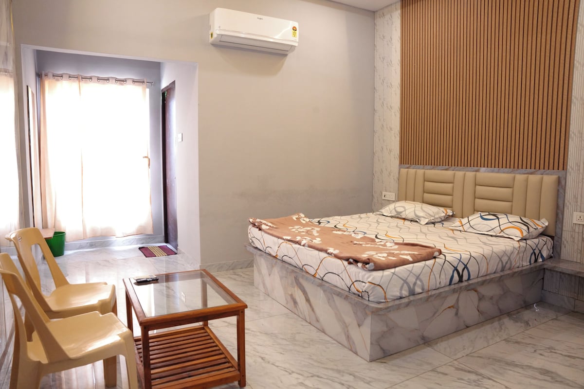 A hotel room with a bed, a glass table, two chairs, a small table, an air conditioning unit, and a curtain.