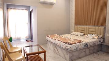 Habitación de hotel con cama, mesa de vidrio, dos sillas, una mesita auxiliar, equipo de aire acondicionado y cortina.