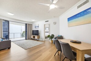 A modern living room with a flat-screen TV, a dining table with chairs, a ceiling fan, and a large window with a view. - 10/21 The Esplanade - A Coastal Escape in the Heart of Cotton Tree (Maroochydore)