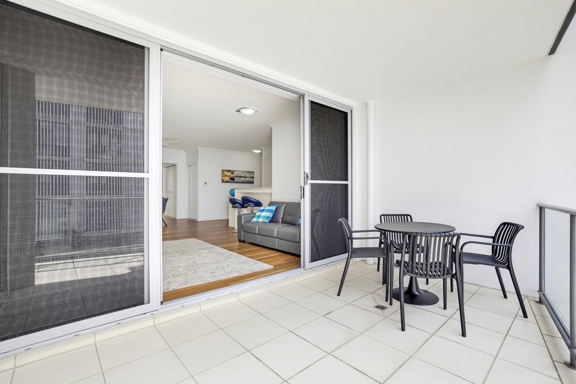 A modern living space with a sliding glass door leading to a balcony, a grey sofa with blue cushions, and a round table with chairs.