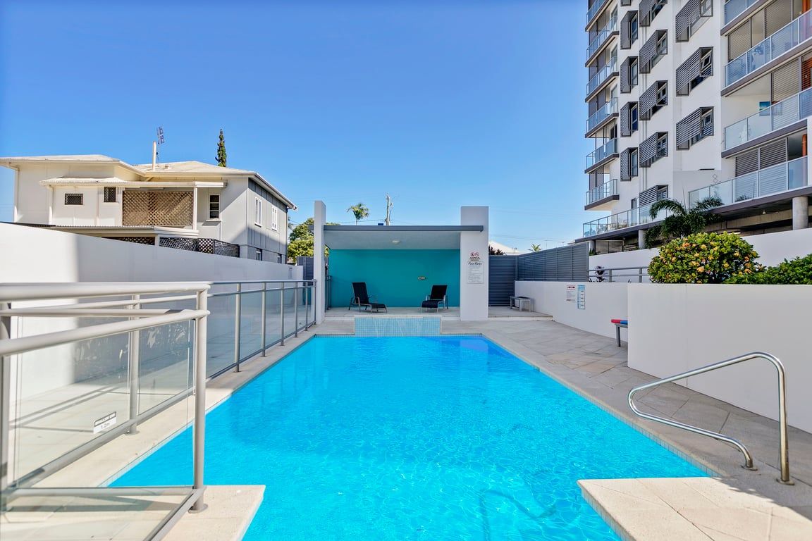 A swimming pool with a clear blue water, surrounded by a white wall and metal railings, with a building in the background.