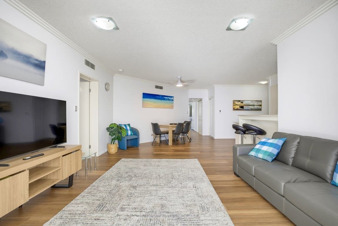 A modern living room with a flat-screen TV, a grey sofa, a dining area with a table and chairs, and a large rug.