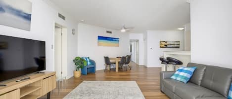 A modern living room with a flat-screen TV, a grey sofa, a dining area with a table and chairs, and a large rug.