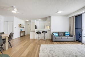 A modern living room with a grey sofa, a dining table with chairs, a kitchen area, and a large window with curtains. - 10/21 The Esplanade - A Coastal Escape in the Heart of Cotton Tree (Maroochydore)