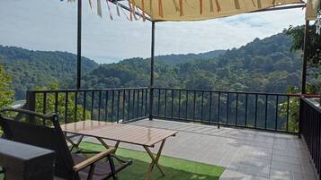 A terrace with a table and chairs, a view of a forested landscape, and a safety railing.