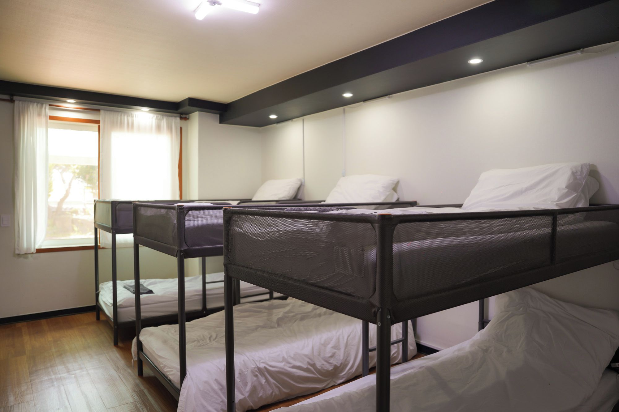 A dormitory room with bunk beds, a window with curtains, and a ceiling light.
