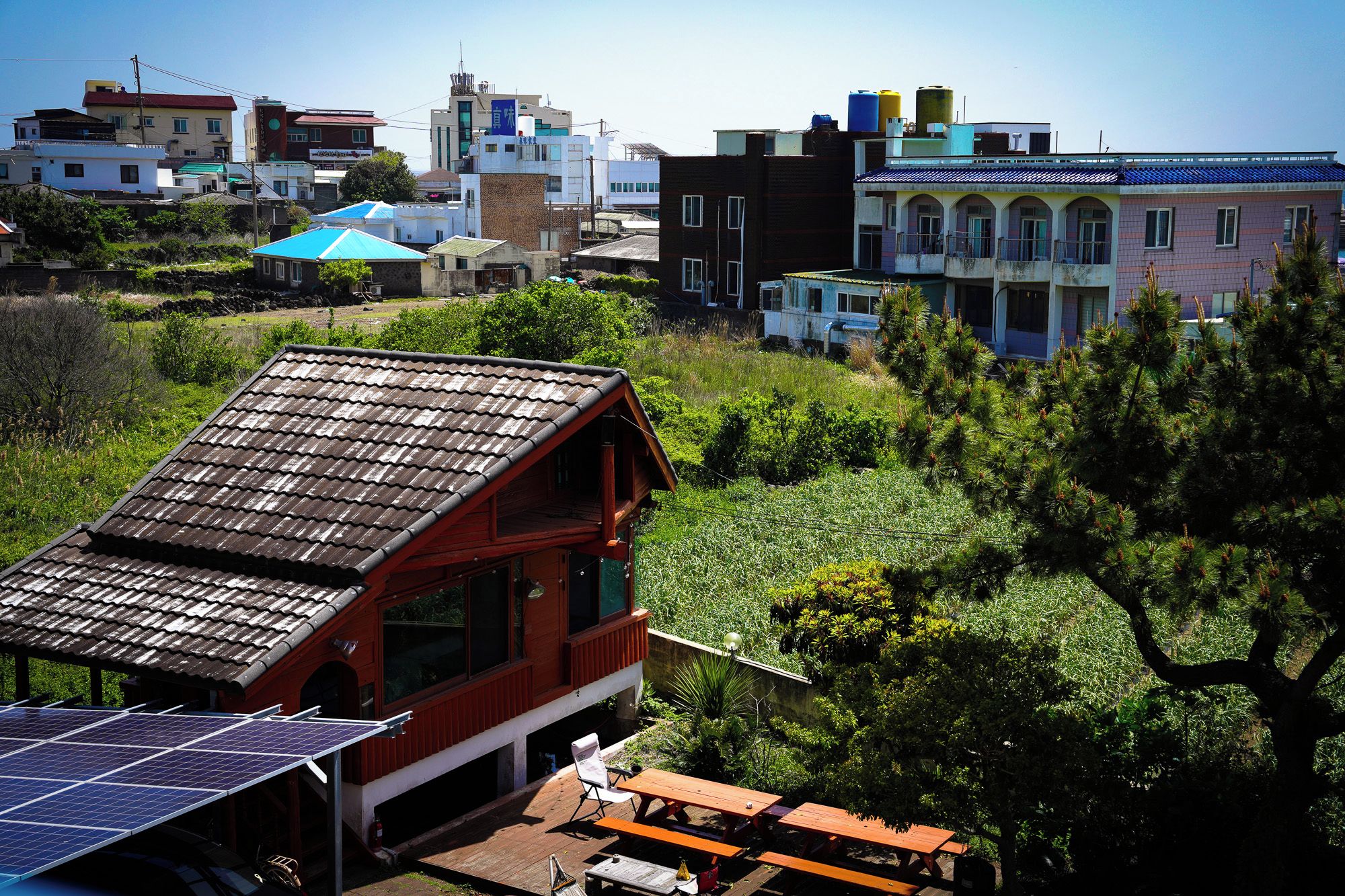 A residential area with a house featuring a sloped roof and solar panels, surrounded by greenery and other buildings.