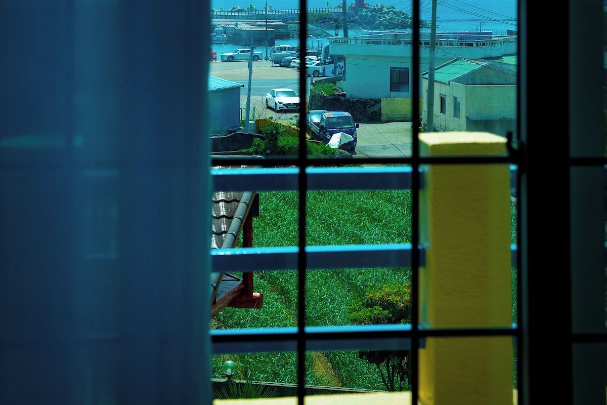 Vista pela janela, com padrão de grade, de uma área costeira com edifícios, um cais e um farol.