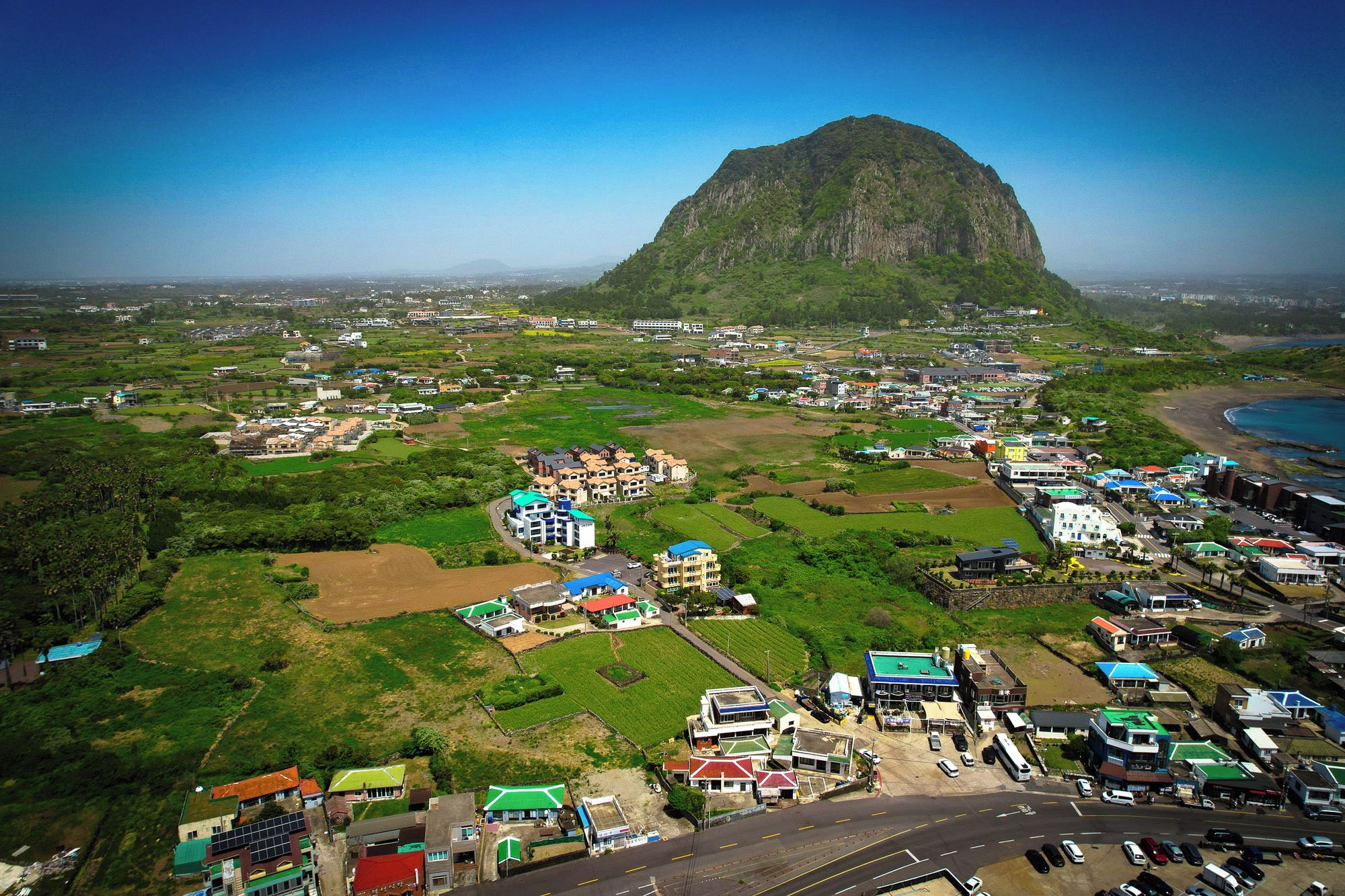 An aerial view of a coastal town with a prominent mountain in the background, a mix of residential and commercial buildings, and a road leading towards the sea.