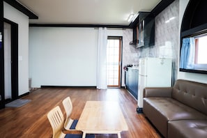 A modern living room with a sofa, a dining table with chairs, a kitchen area with a refrigerator, and a sliding glass door leading to a balcony.