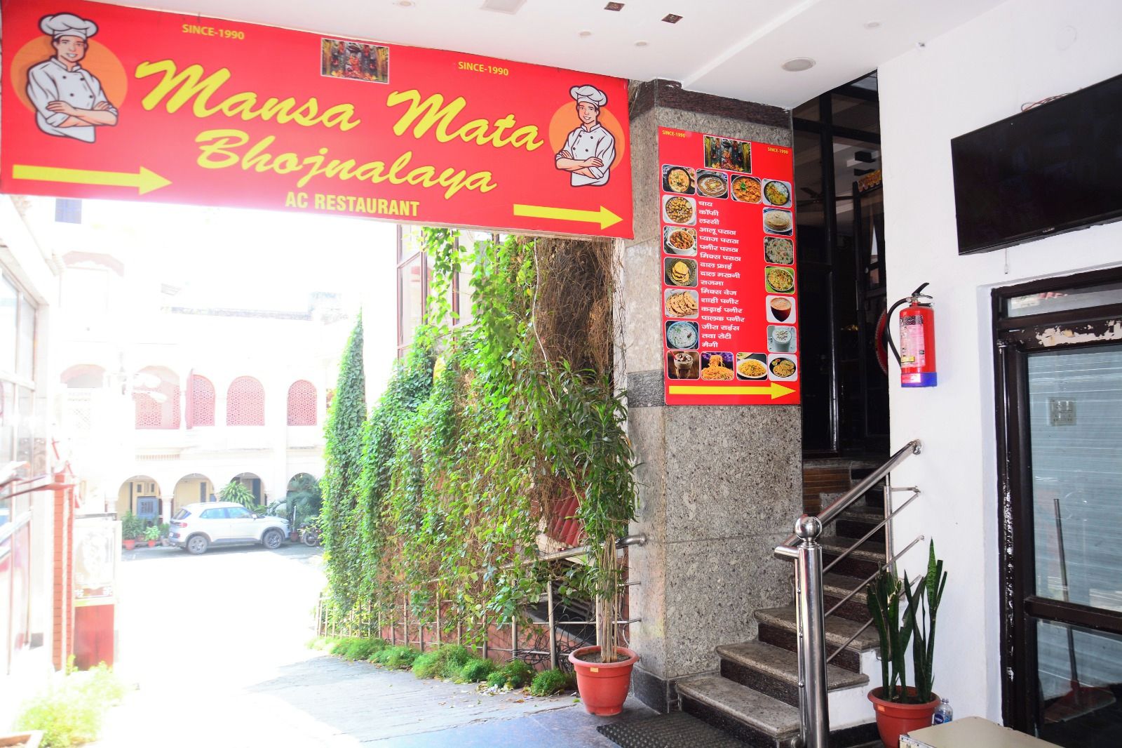 The entrance to Mansa Mata Bhujanalaya AC Restaurant with a staircase leading up to the entrance, a fire extinguisher mounted on the wall, and a menu board displaying various dishes.