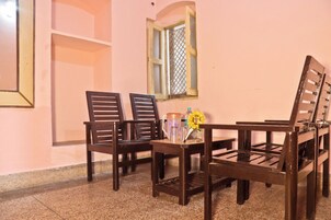 Una habitación con un banco de madera, dos sillas, una mesa pequeña con un jarrón de flores amarillas y una ventana con mosquitero.