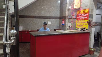 A reception desk with a man seated behind it, a fire extinguisher mounted on the wall, and a sign indicating check-out time and mandatory GST.