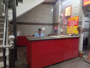 A reception desk with a man seated behind it, a fire extinguisher mounted on the wall, and a sign indicating check-out time and mandatory GST.