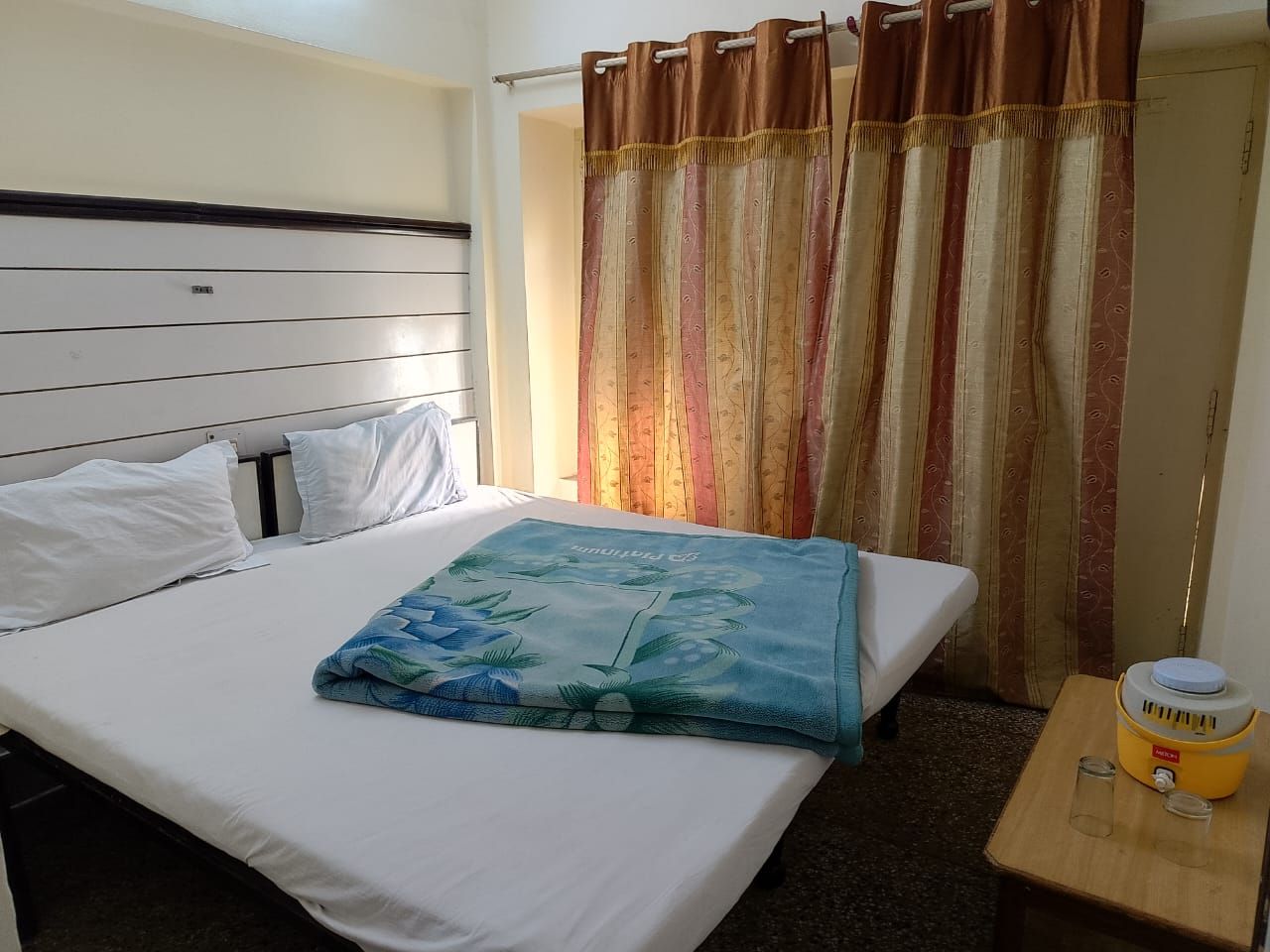 A hotel room with a bed, a bedside table with a water dispenser, and a curtain.