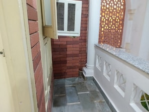 A narrow outdoor corridor with a brick wall, a white door, a window with blinds, and a white balustrade.