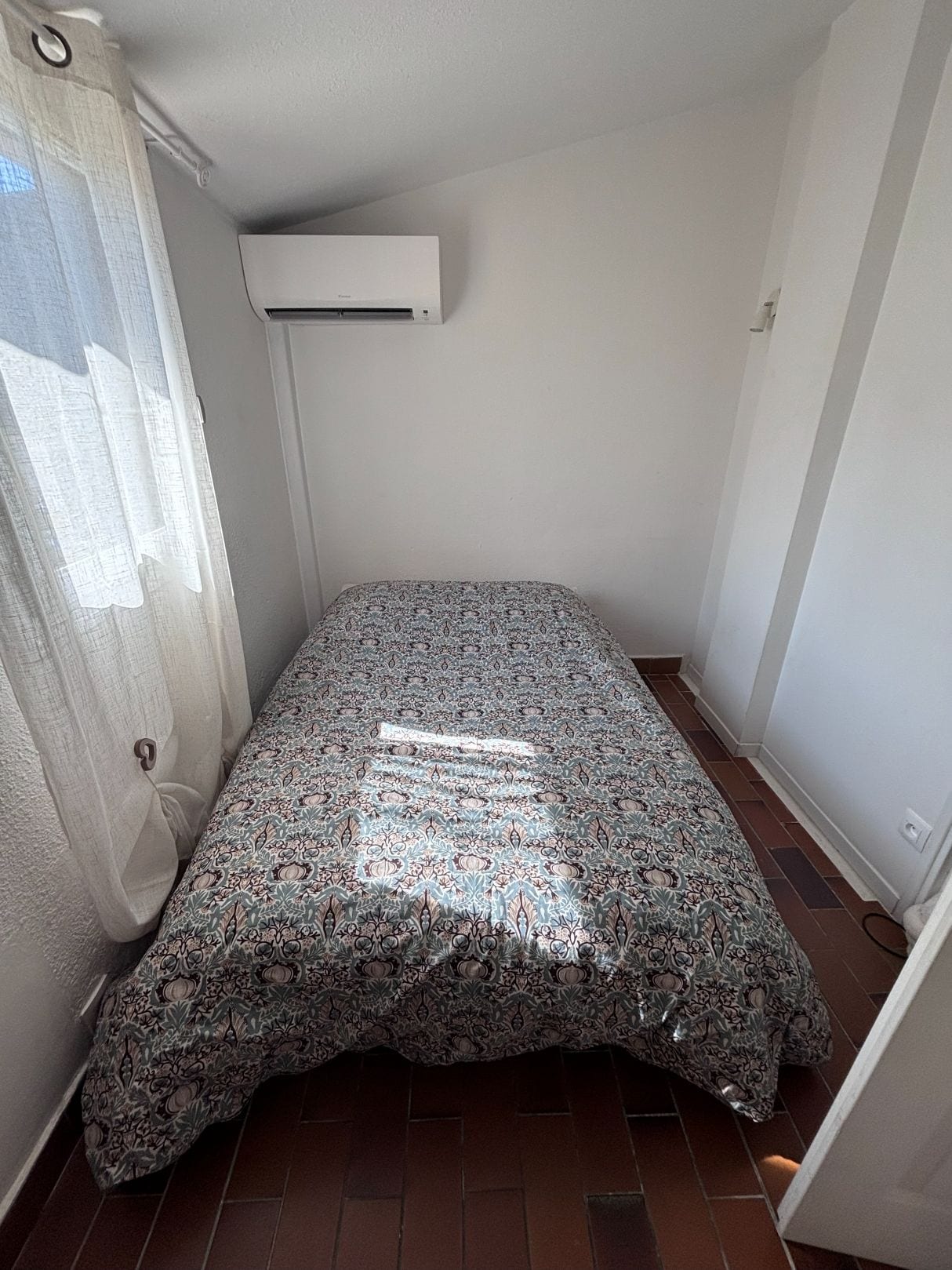 A small room with a patterned bedspread, a window with curtains, and an air conditioning unit on the wall.