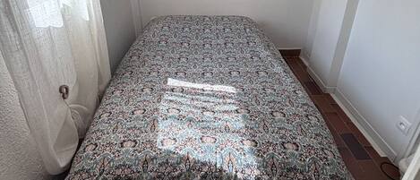 A small room with a patterned bedspread, a window with curtains, and an air conditioning unit on the wall.