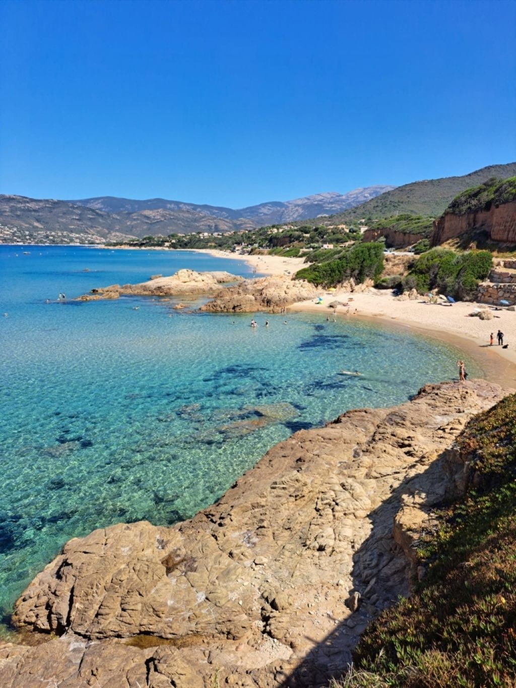 A coastal landscape with clear blue waters, rocky shores, and a sandy beach.