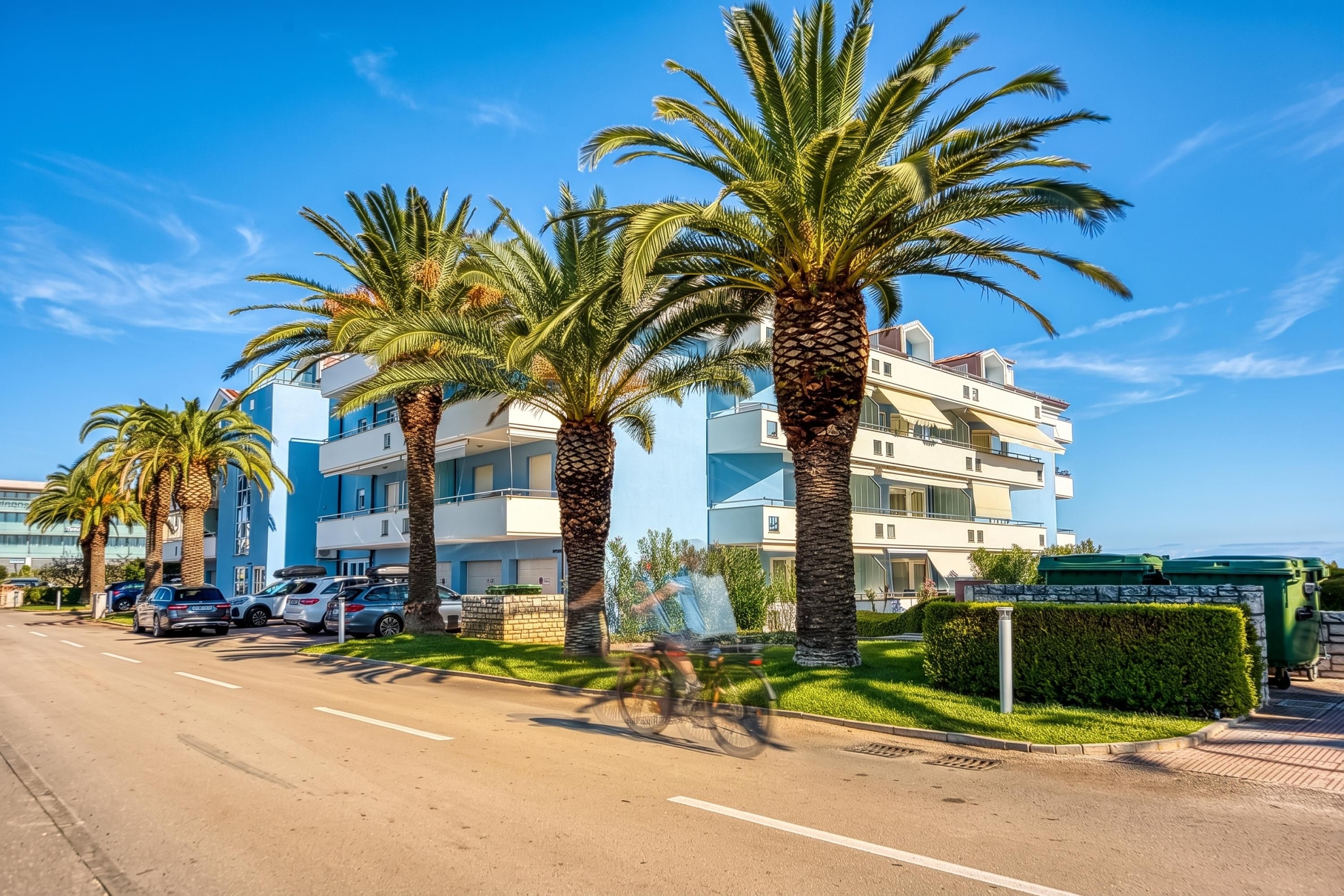 Un immeuble à plusieurs étages doté de balcons, avec des voitures garées et des palmiers.