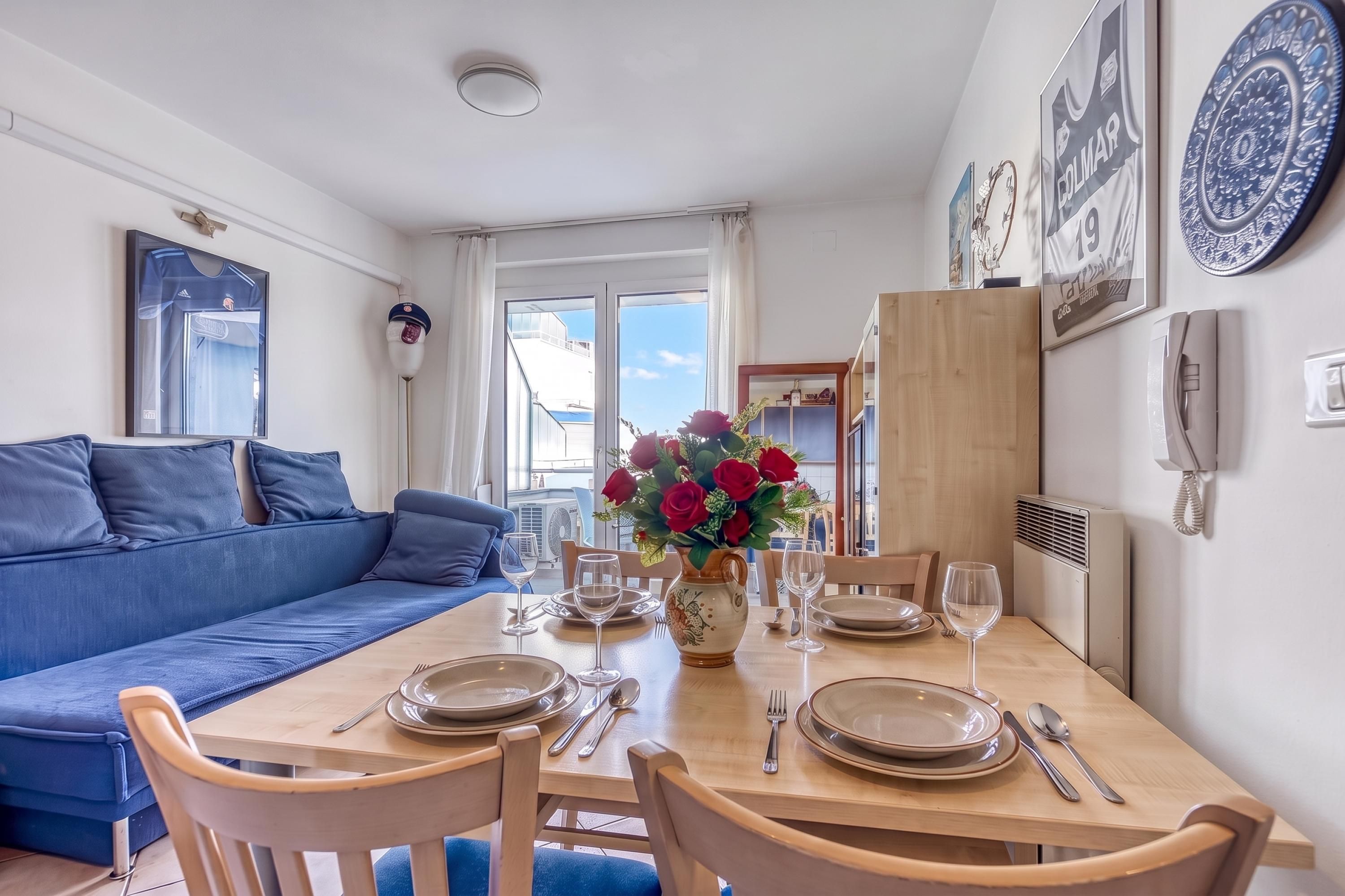 Een eethoek met een houten tafel voor vier personen, een blauwe bank en een vaas met rode bloemen.