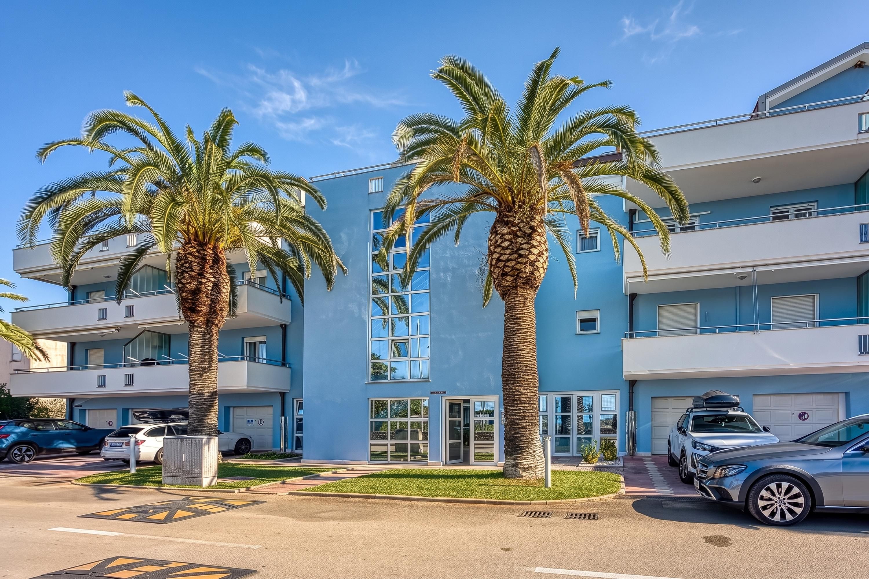 Un immeuble moderne à plusieurs étages, doté d’une façade bleue, bordé de palmiers et avec des voitures garées dans l’allée.