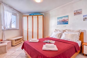 A hotel room with a bed, a wooden nightstand, a bench, a window with curtains, a wardrobe, and a wall with two paintings. - Residence Skiper Savudrija Apt 153 Pool & Gym (Umag)