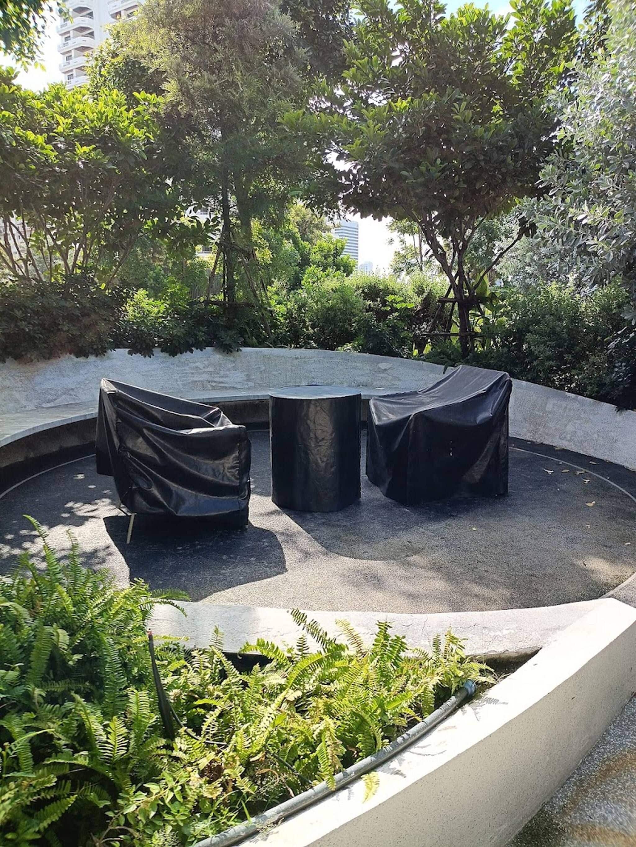 An outdoor seating area with a circular arrangement of tables and chairs, surrounded by greenery and a concrete wall.