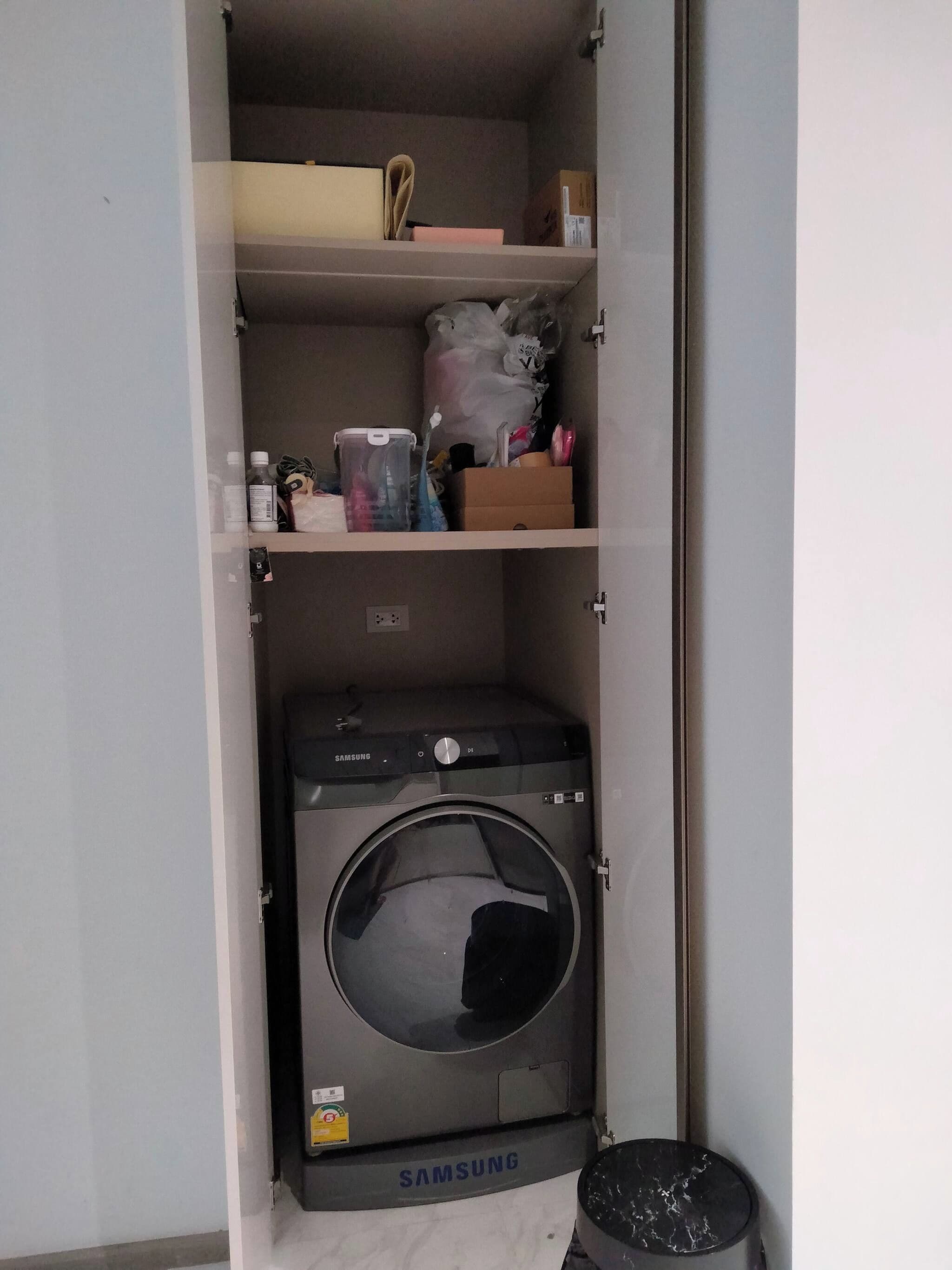 A laundry room with a Samsung washing machine, shelves with various items, and a trash bin.