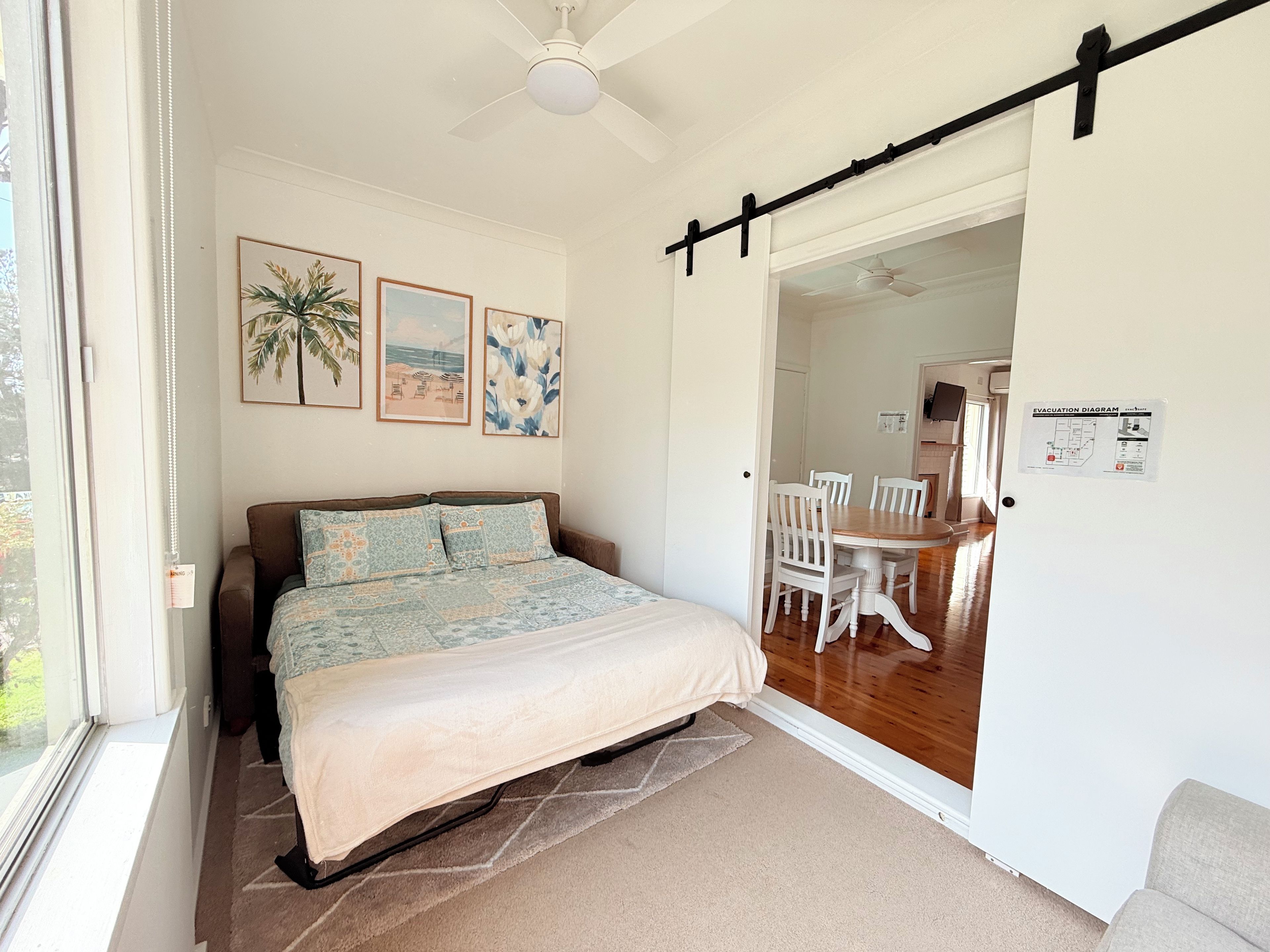 A bedroom with a bed, a ceiling fan, a dining area with chairs and a table, and a door with an evacuation diagram.