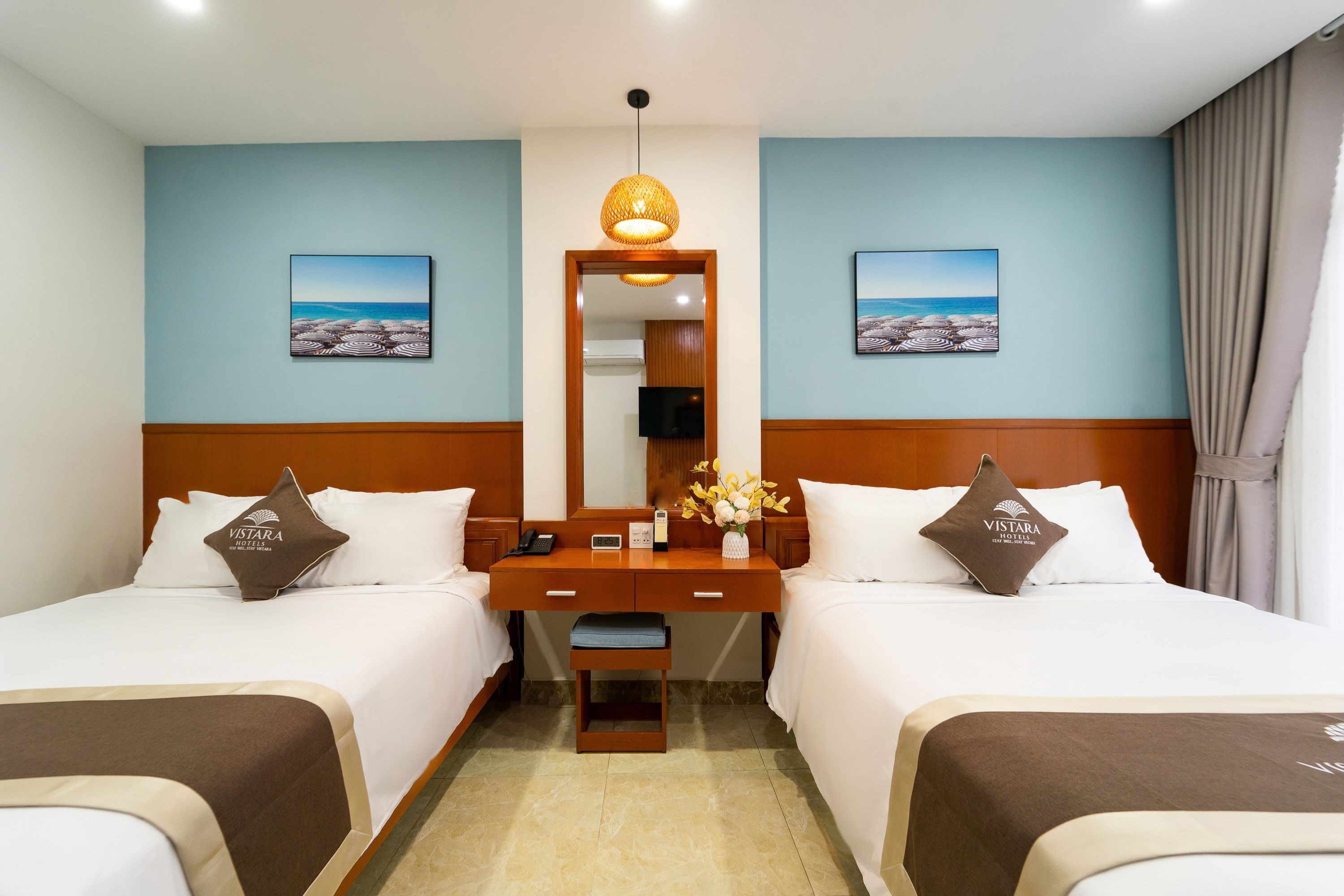Habitación de hotel con dos camas, una mesita de noche de madera, un espejo y dos cuadros con imágenes de playas.