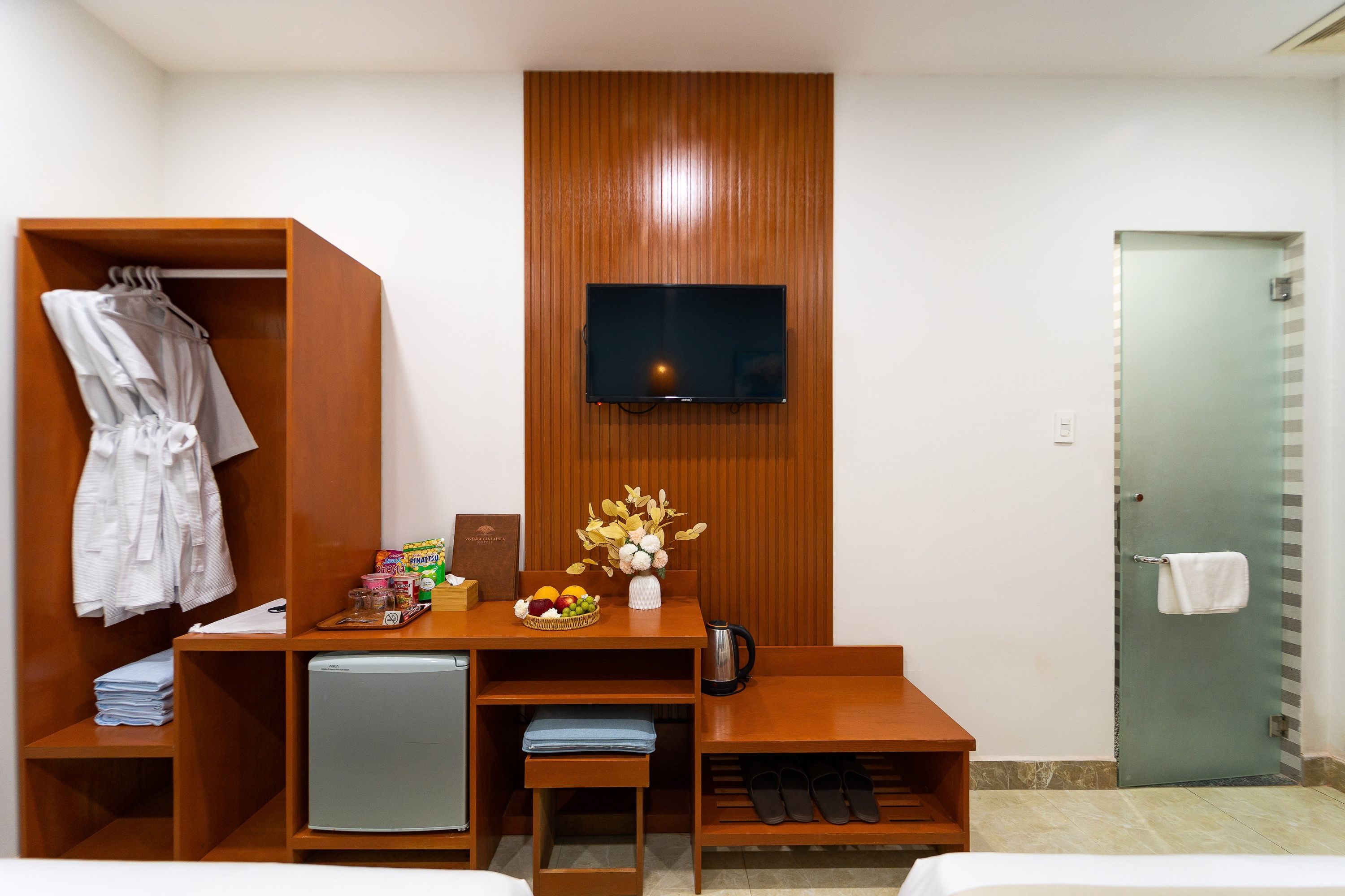 Quarto de hotel com uma escrivaninha de madeira, televisão presa na parede, guarda-roupa, pequena mesa com cesta de frutas e uma porta com uma toalha pendurada.