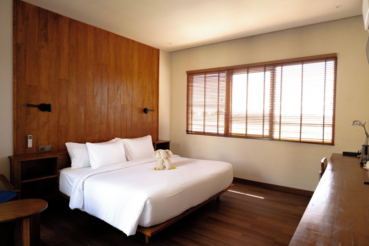 A bedroom with a wooden headboard, a large bed with white bedding, a bedside table, and a window with blinds.