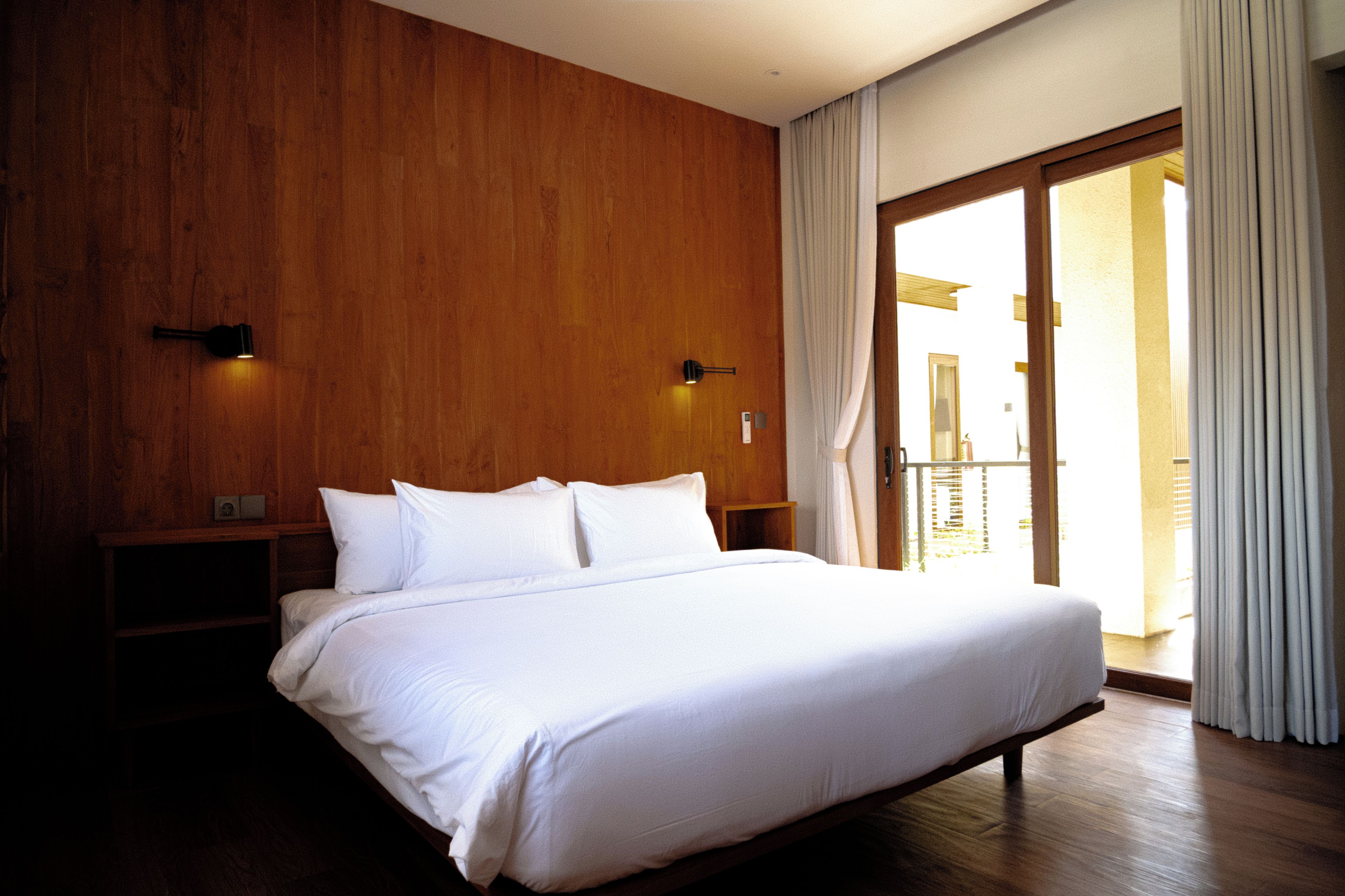 Habitación de hotel con pared de madera, cama con ropa blanca, mesita de noche, lámpara de pared y puerta corrediza de vidrio que da a un espacio al aire libre.