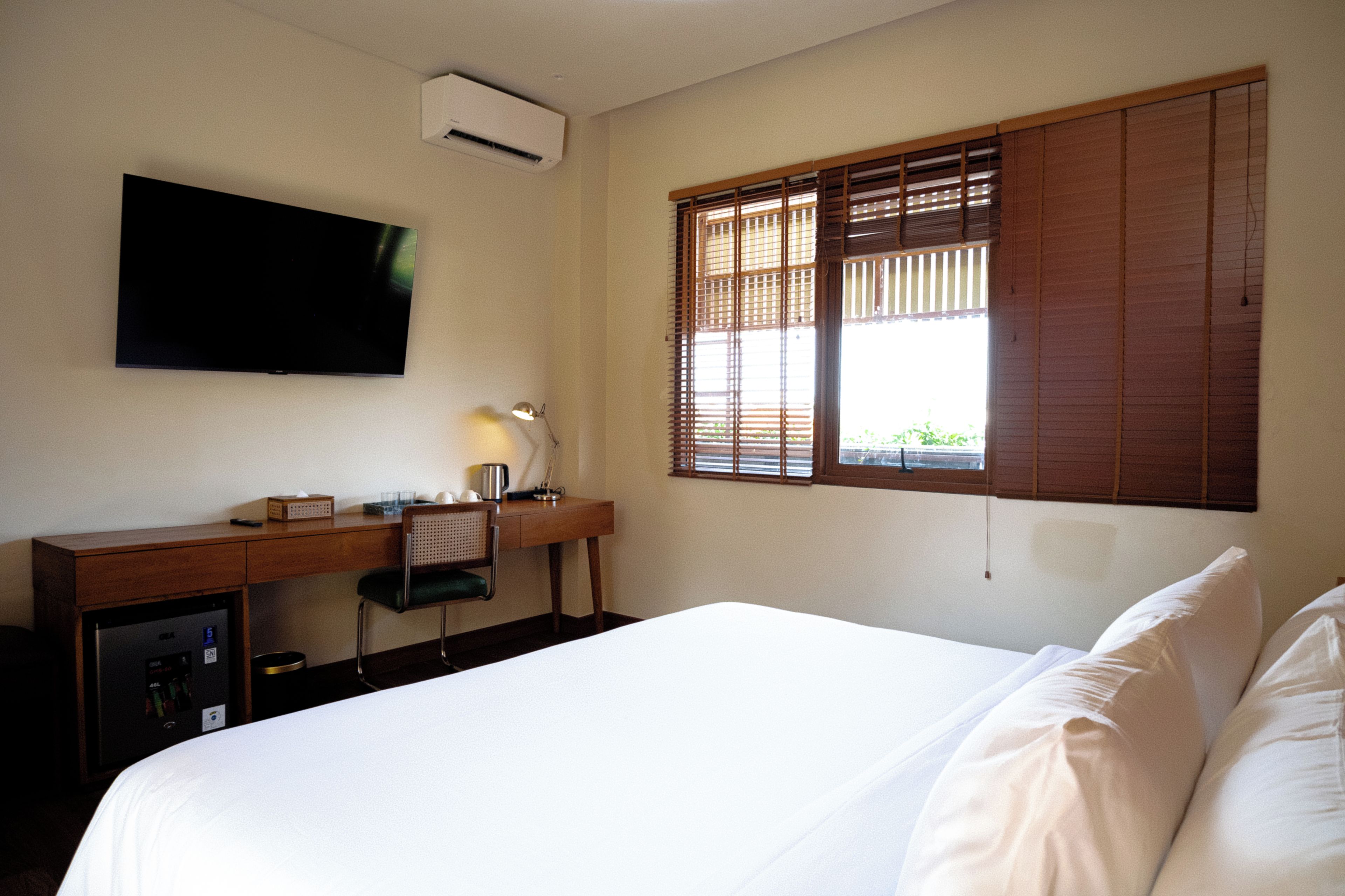 A hotel room with a bed, a desk with a lamp, a television, and a window with blinds.